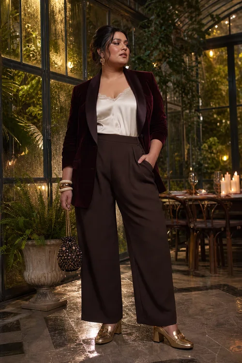 A woman wears a burgundy velvet blazer with an ivory camisole, brown wide-leg trousers, metallic gold heeled loafers, bangles, earrings, and a small beaded handbag.
