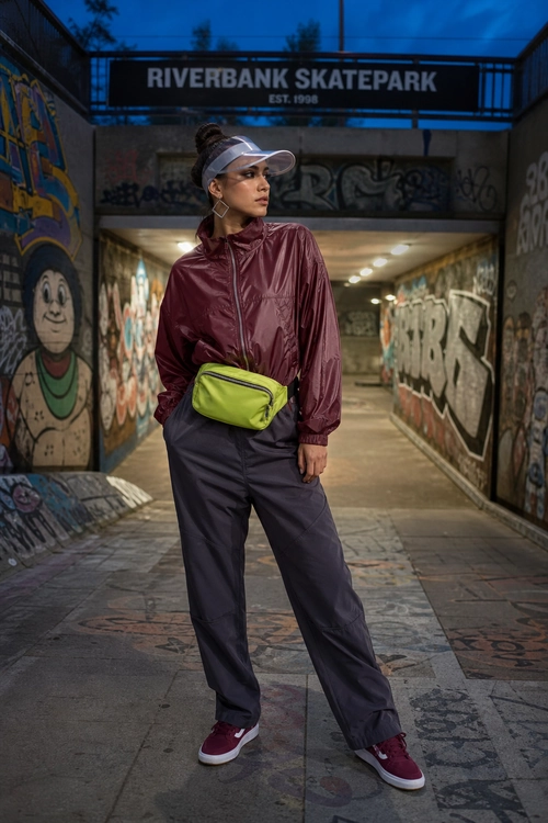A sporty streetwear outfit featuring a burgundy zip-up windbreaker, dark loose-fit pants, burgundy sneakers, a lime green waist bag, and a translucent visor.