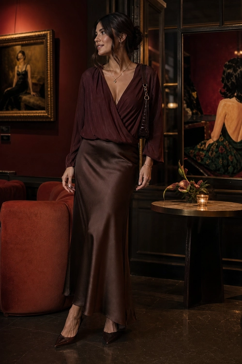 A woman wears a burgundy draped wrap top with a brown satin maxi skirt, pointed heels, and a small chain-strap shoulder bag.