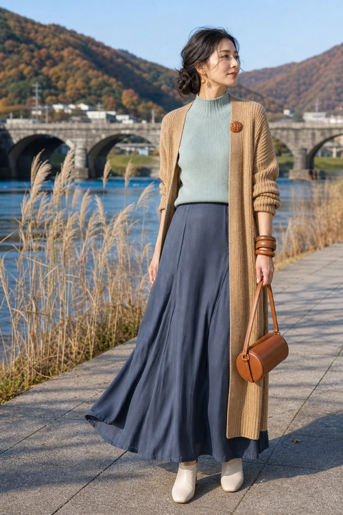 Camel Cardigan and Navy Skirt Outfit Woman wearing a long camel cardigan over a pale green ribbed sweater with a flowing navy skirt, cream ankle boots, and a brown handbag.