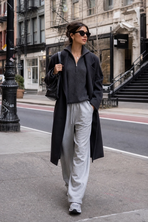 A woman wears a long black coat over a charcoal quarter-zip top with gray wide-leg sweatpants, gray sneakers, black sunglasses, and a black shoulder bag.