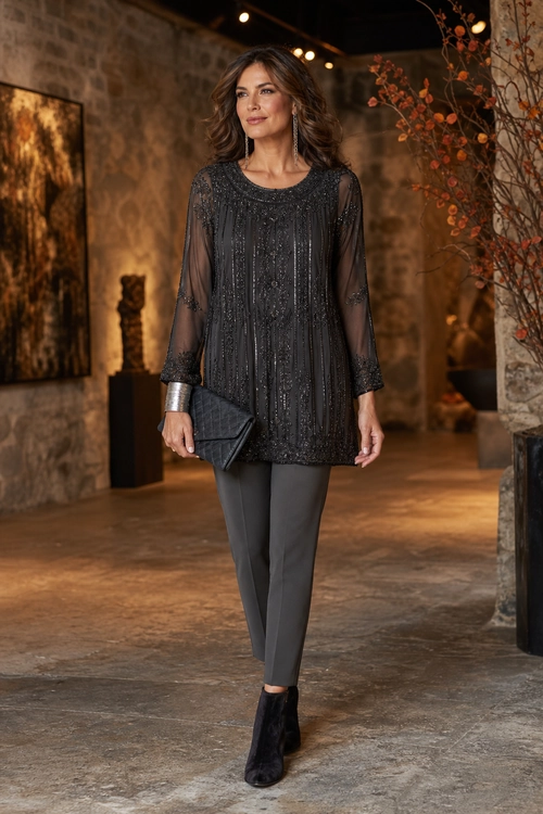 A woman wears a charcoal embellished tunic with sheer sleeves, gray slim trousers, black ankle boots, and a black clutch.