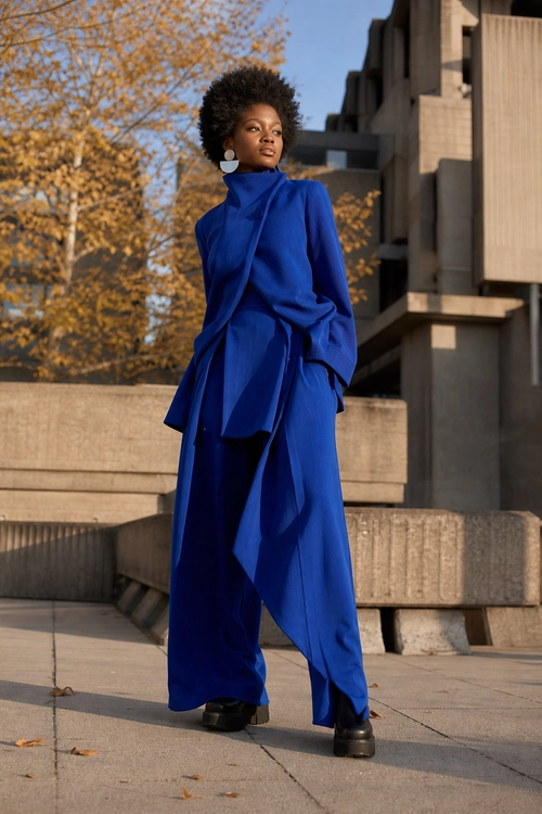 A person wears a cobalt blue high-neck draped top with wide-leg trousers, black shoes, and white statement earrings outdoors.