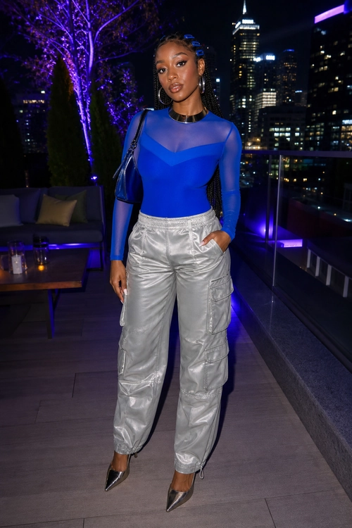 Cobalt Blue Top with Silver Cargo Pants A woman wears a cobalt blue sheer-panel long-sleeve top with silver cargo pants, pointed black heels, and a black shoulder bag at night.