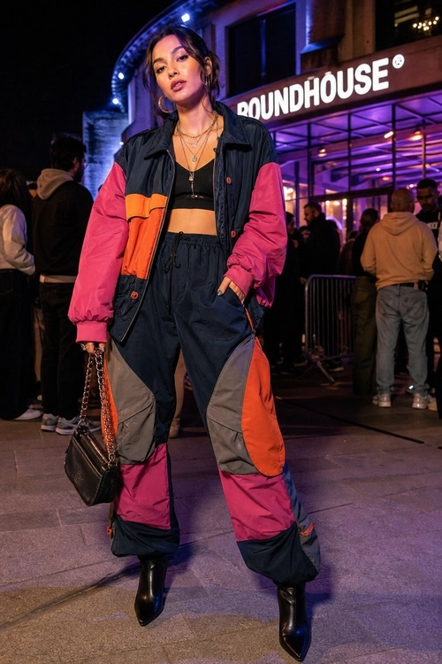 A woman wears a color-blocked bomber jacket with a black crop top, oversized utility pants, black ankle boots, and a chain-strap handbag.