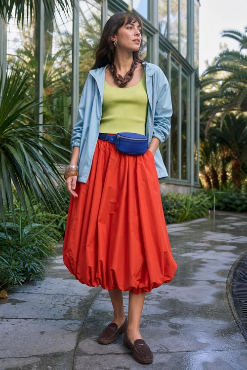 Woman wearing a light blue jacket, lime green sleeveless top, red-orange midi skirt, blue waist bag, and brown loafers.