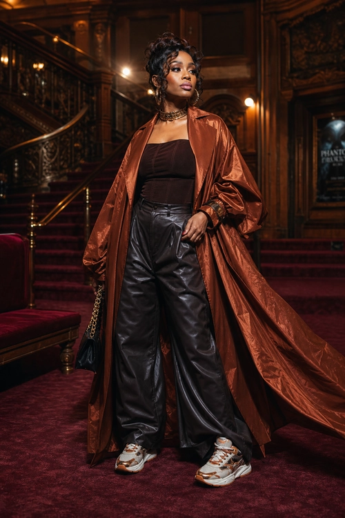 Copper Coat and Black Leather Trousers A woman wears a long copper satin coat over a black strapless top and wide-leg leather trousers with white sneakers.