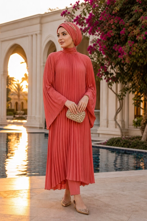 Coral Pleated Modest Outfit A woman wears a coral pleated long dress with a matching wrapped headscarf, ankle-length trousers, embellished clutch, and pointed shoes.