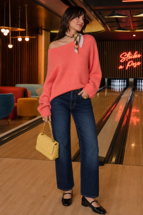 A coral off-shoulder sweater is styled with a patterned neck scarf, dark wide-leg jeans, black Mary Jane shoes, and a pale yellow quilted handbag.