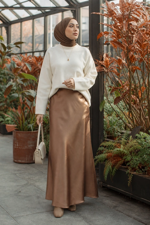 Cream Sweater and Satin Maxi Skirt A modest outfit featuring a cream sweater, brown satin maxi skirt, brown hijab, beige shoes, and a small cream handbag in a greenhouse setting.