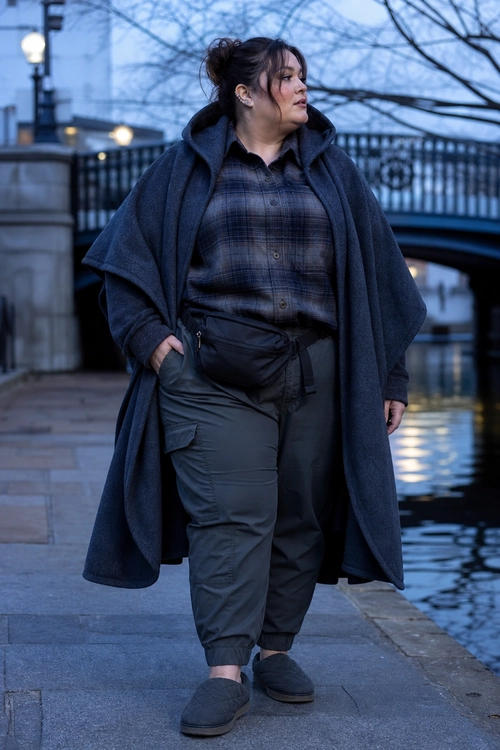 A dark layered outfit featuring a hooded cape coat, plaid button-up shirt, cargo jogger pants, waist bag, and slip-on shoes.