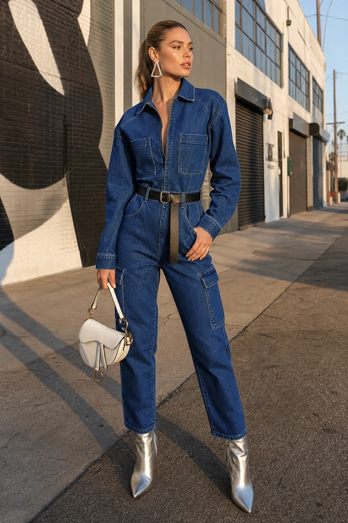 A woman wears a belted blue denim jumpsuit with silver pointed ankle boots, geometric earrings, and a small white handbag.