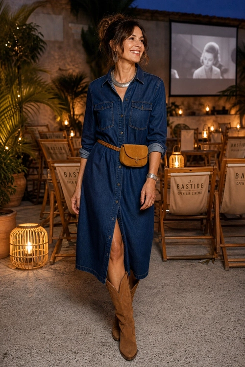 A woman wears a dark blue denim midi shirt dress with a tan waist pouch belt, silver jewelry, and tan knee-high boots.