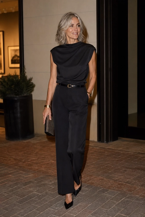 A woman wears a sleeveless black draped top with wide-leg black trousers, a slim belt, pointed pumps, gold jewelry, and a small clutch.