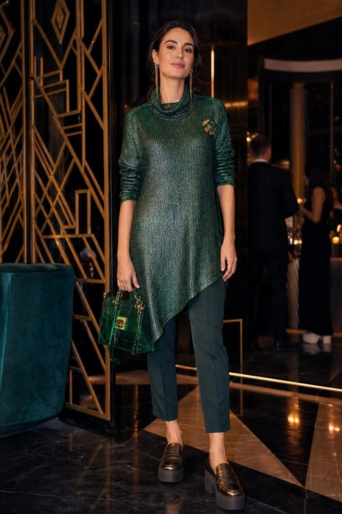 A woman wears a dark green asymmetric tunic with slim trousers, loafers, and a small transparent green handbag.