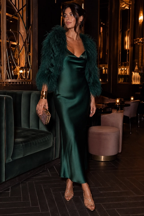 Woman wearing a dark emerald satin slip dress with a feather-textured jacket, strappy heels, and gold accessories.