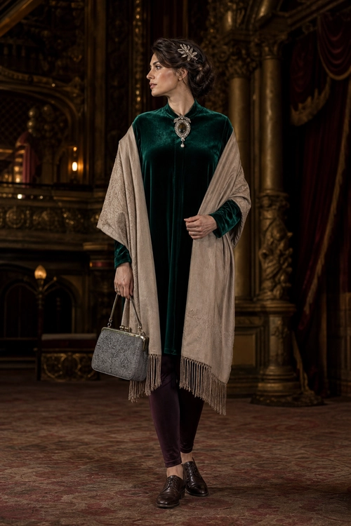 A woman wears a dark green velvet dress with a beige fringed shawl, burgundy tights, black lace-up shoes, and a gray handbag.