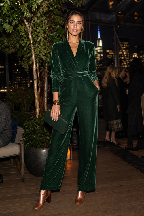 A woman wears a dark green velvet wrap jumpsuit with wide legs, copper ankle boots, and a matching dark clutch.