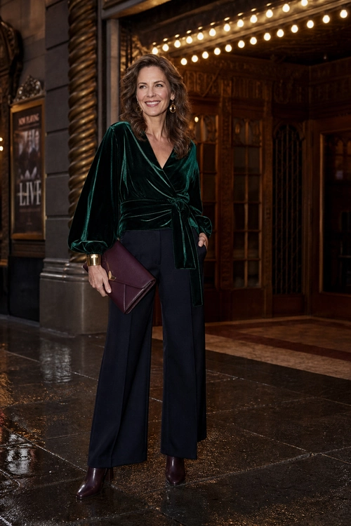 A woman wears an emerald green velvet wrap top with navy wide-leg trousers, burgundy ankle boots, and a matching burgundy clutch.