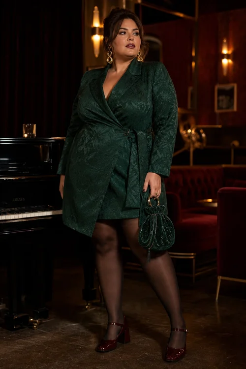 A woman wears a dark emerald wrap dress with sheer black tights, burgundy heels, gold earrings, and a matching green handbag.