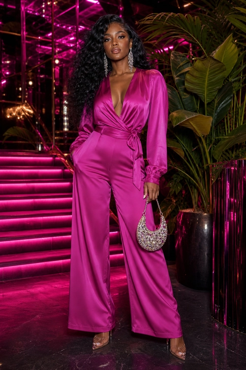 Fuchsia Satin Jumpsuit Evening Look A woman wears a vivid fuchsia satin wrap-front jumpsuit with wide legs, strappy heels, dangling earrings, and a silver embellished crescent handbag.