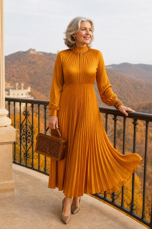 Golden Pleated Dress A woman wears a long golden pleated dress with a high neckline, pointed heels, and a structured brown handbag on a balcony.