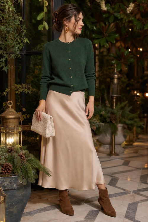 A dark green button-front cardigan is paired with a champagne satin midi skirt, brown ankle boots, and a woven beige clutch.