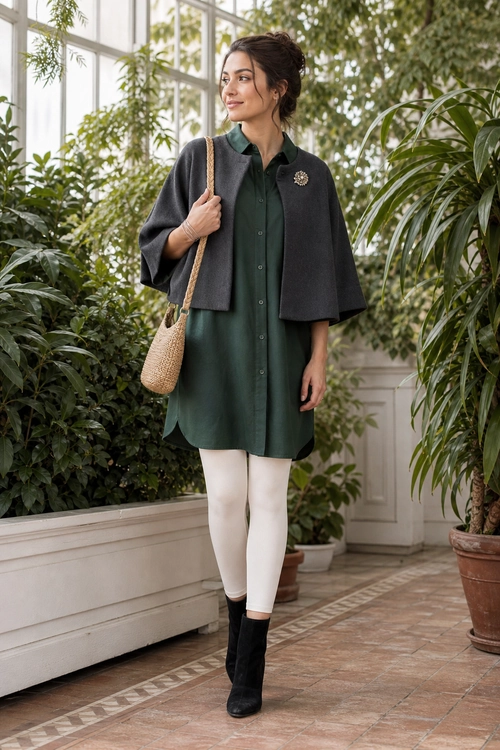 A dark green button-front tunic dress is layered with a gray cape, cream tights, black ankle boots, and a woven shoulder bag.