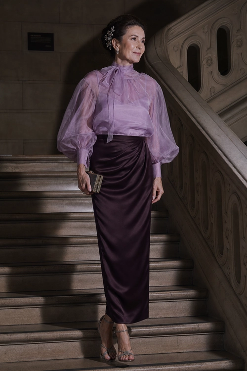 A woman wears a sheer lavender high-neck blouse with voluminous sleeves, a plum satin skirt, strappy heels, and a small metallic clutch.