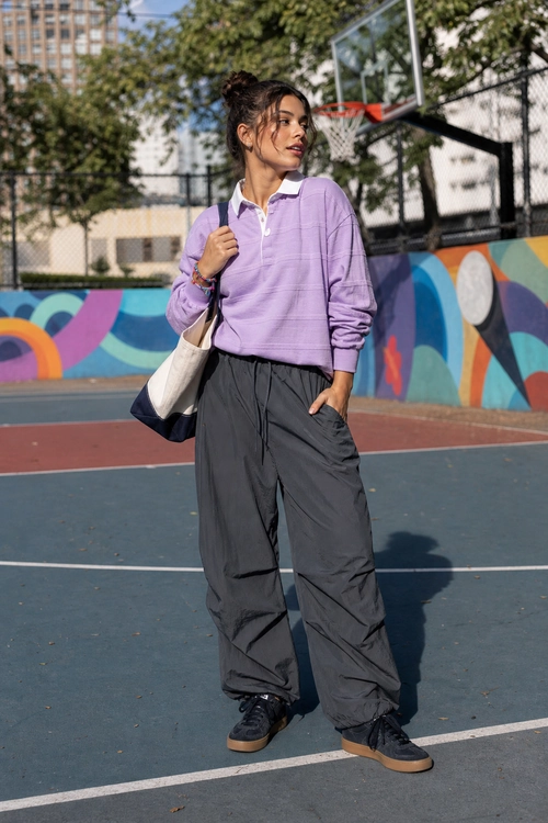 A lavender collared pullover with charcoal wide-leg pants, black sneakers, and a two-tone tote bag worn on an outdoor basketball court.