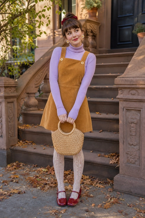 A lavender turtleneck layered under a mustard corduroy pinafore dress with patterned tights, red Mary Jane shoes, and a round woven handbag.
