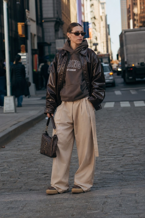 Street-style outfit featuring a dark leather jacket over a hooded sweatshirt, beige wide-leg trousers, brown shoes, sunglasses, and a patterned handbag.