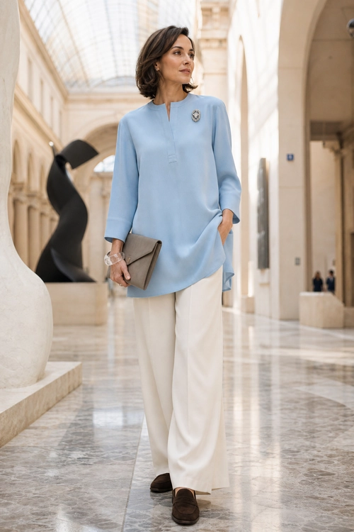 A woman wears a light blue tunic blouse with cream wide-leg trousers, brown loafers, and a taupe clutch.