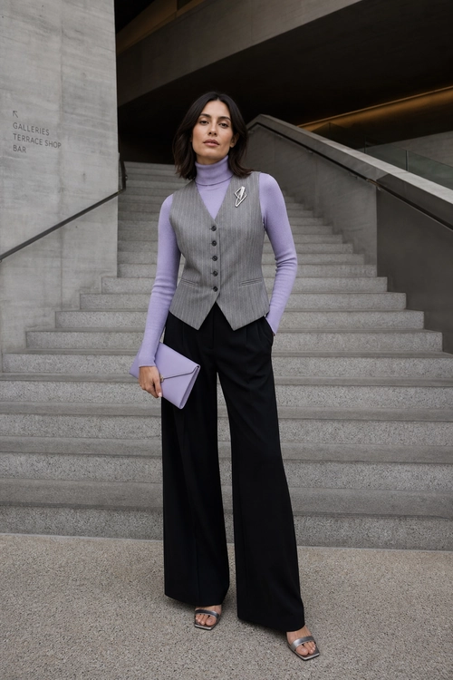 Lilac Turtleneck with Tailored Vest A woman wears a lilac turtleneck, gray pinstriped buttoned vest, black wide-leg trousers, a matching clutch, and strappy sandals on a staircase.