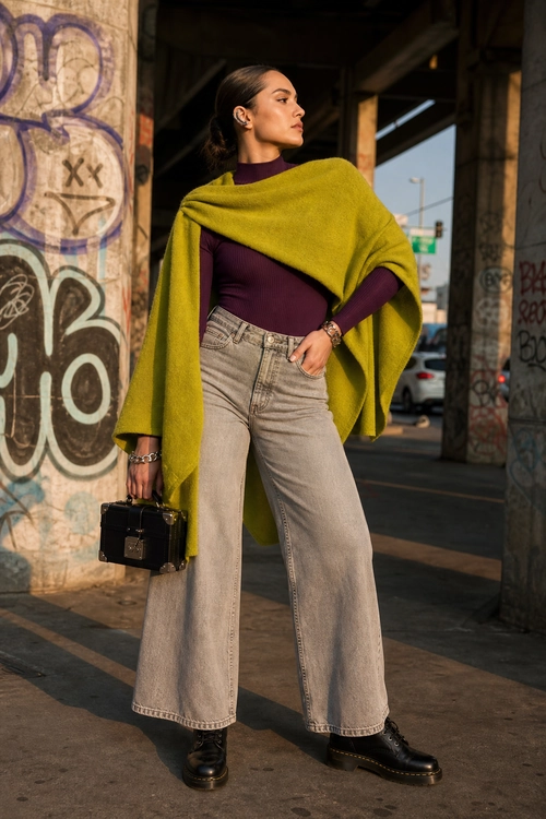 A woman wears a lime green draped cape over a purple turtleneck with high-waisted wide-leg jeans, black boots, and a structured black handbag.