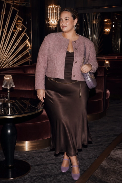 A mauve cropped jacket styled over a dark brown satin dress with a chain belt, lavender shoulder bag, and matching Mary Jane heels.