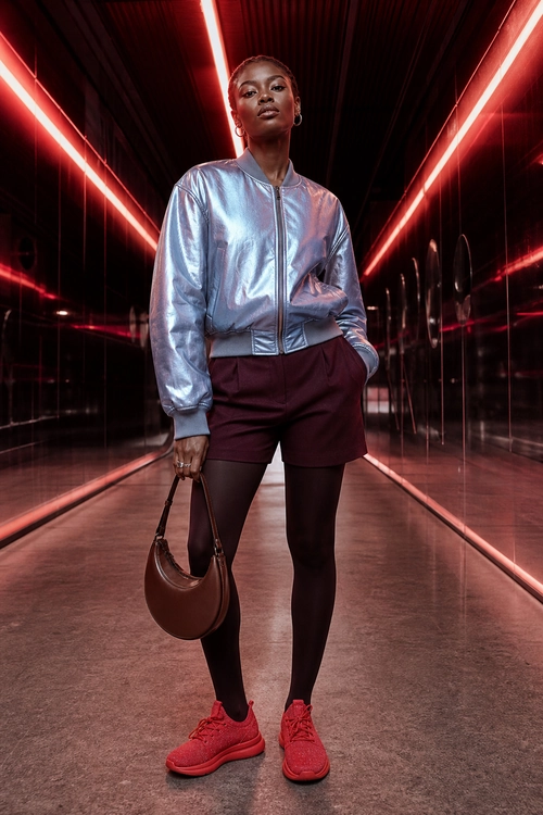 Metallic Bomber Jacket with Burgundy Shorts Fashion outfit featuring a metallic silver bomber jacket, burgundy shorts, black tights, red sneakers, and a brown shoulder bag.