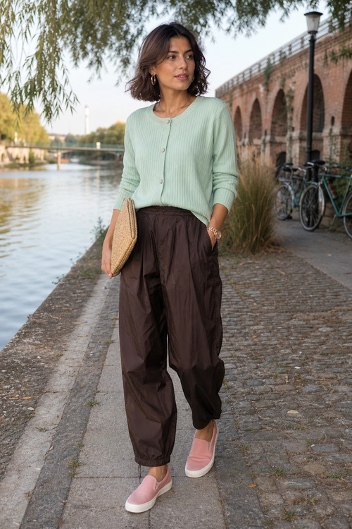 Woman wearing a mint green button-front cardigan, dark brown jogger pants, pink slip-on sneakers, and carrying a woven beige clutch.