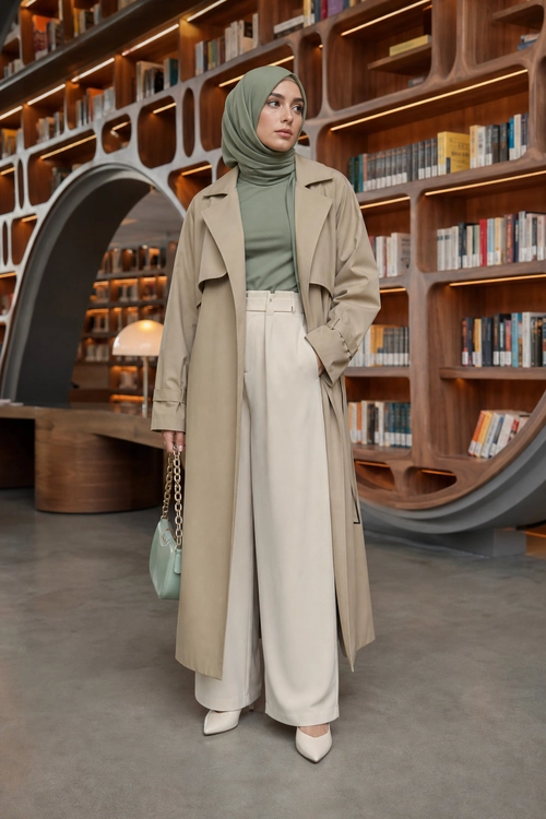 Modest Neutral Layered Outfit A woman wears a beige long trench coat over a muted green top, cream wide-leg trousers, a matching hijab, white pointed shoes, and a small mint handbag.
