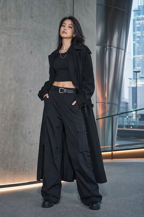 A woman wears a black cropped top, long black coat, wide-leg cargo trousers, belt, necklace, and black shoes in a modern city interior.