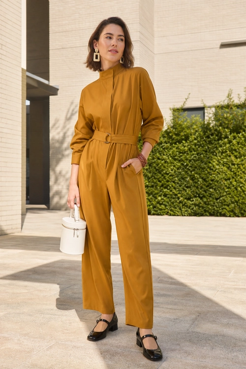 Mustard Belted Jumpsuit Outfit Woman wearing a mustard belted jumpsuit with black Mary Jane shoes, a white bucket bag, and geometric earrings.
