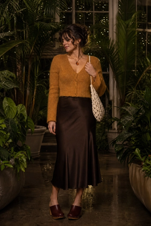 A woman wears a mustard cropped cardigan with a dark brown satin midi skirt, burgundy clogs, a cream crochet shoulder bag, and a pendant necklace.