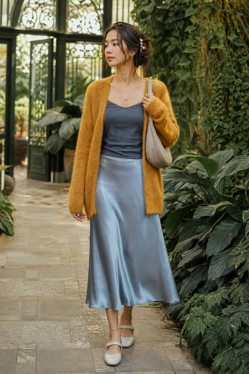 A woman wears a mustard cardigan over a dark blue camisole with a blue-gray satin midi skirt, cream Mary Jane shoes, and a beige shoulder bag.