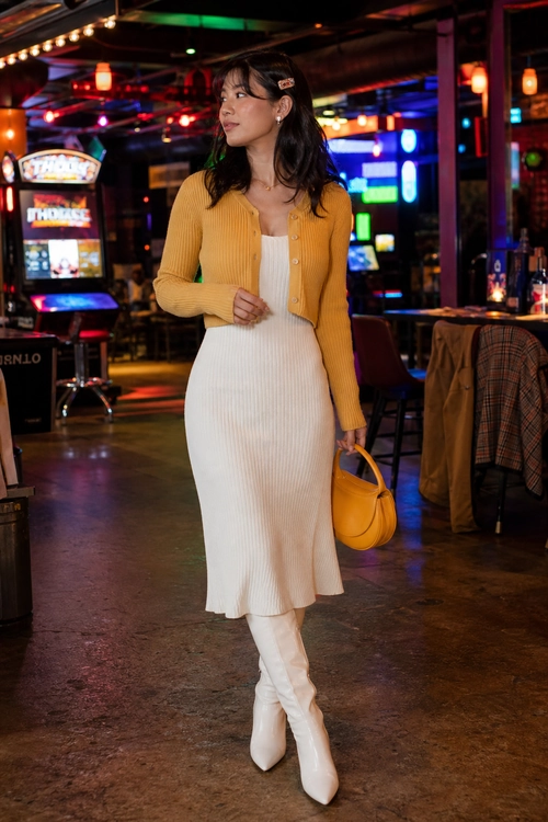 A woman wears a mustard cardigan over a white ribbed midi dress with white knee-high boots and a matching mustard handbag.
