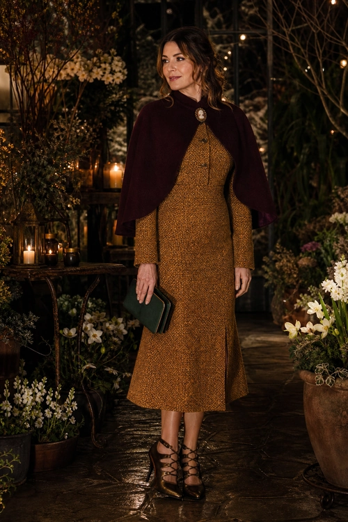 A woman wears a textured mustard midi dress with a burgundy cape, dark clutch, and lace-up heeled sandals.