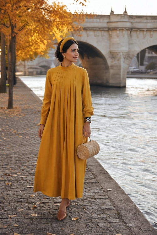 A woman wears a long mustard pleated dress with a matching headband, brown shoes, and a woven cylindrical handbag beside a riverside bridge.