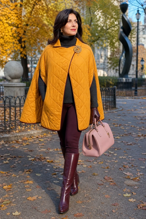 A woman wears a mustard quilted cape over a black turtleneck with burgundy trousers, knee-high boots, and a pink handbag.
