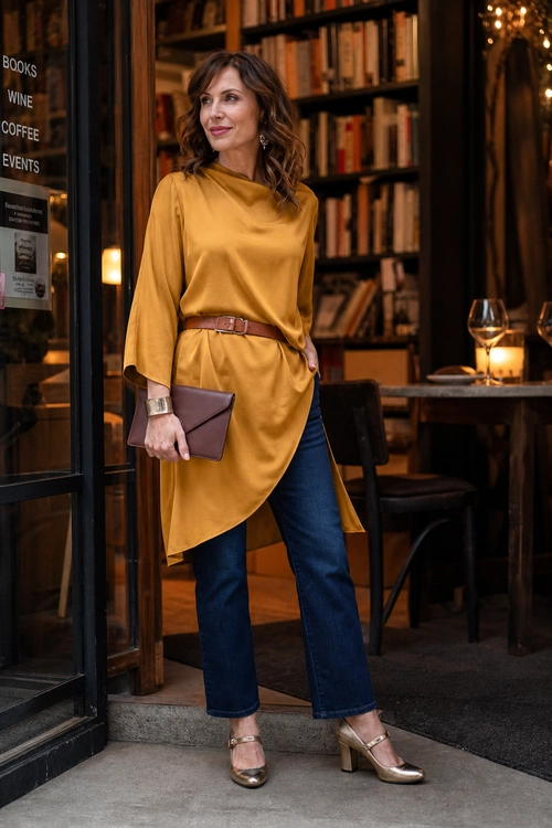 Woman wearing a mustard asymmetrical tunic belted at the waist with blue jeans, metallic heels, a brown clutch, cuff bracelet, and drop earrings.