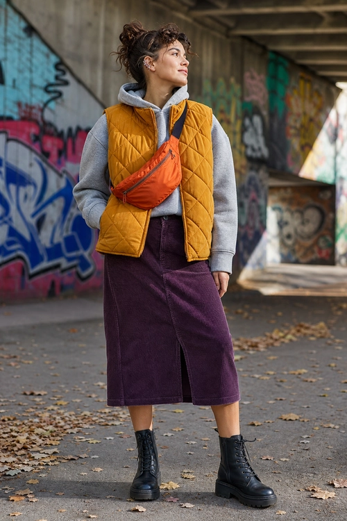 A casual outfit featuring a gray hoodie, mustard quilted vest, burgundy corduroy midi skirt, orange crossbody bag, and black lace-up boots.