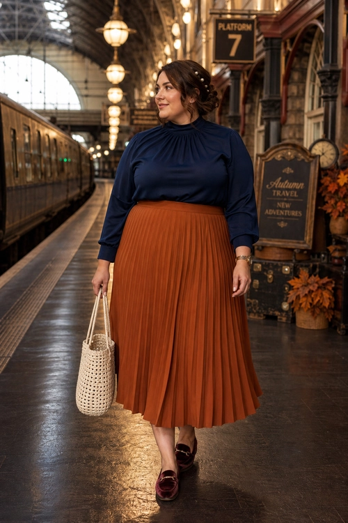 Navy Top and Rust Pleated Skirt Outfit A woman wears a navy long-sleeve top with a rust pleated midi skirt, burgundy loafers, and a woven tote at a train station.